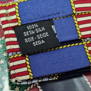 May include: Close-up of a silk tie with a colorful geometric pattern. The tie features blue, red, and white stripes, with yellow rope-like borders. A black label indicates the tie is 100% seta-silk.