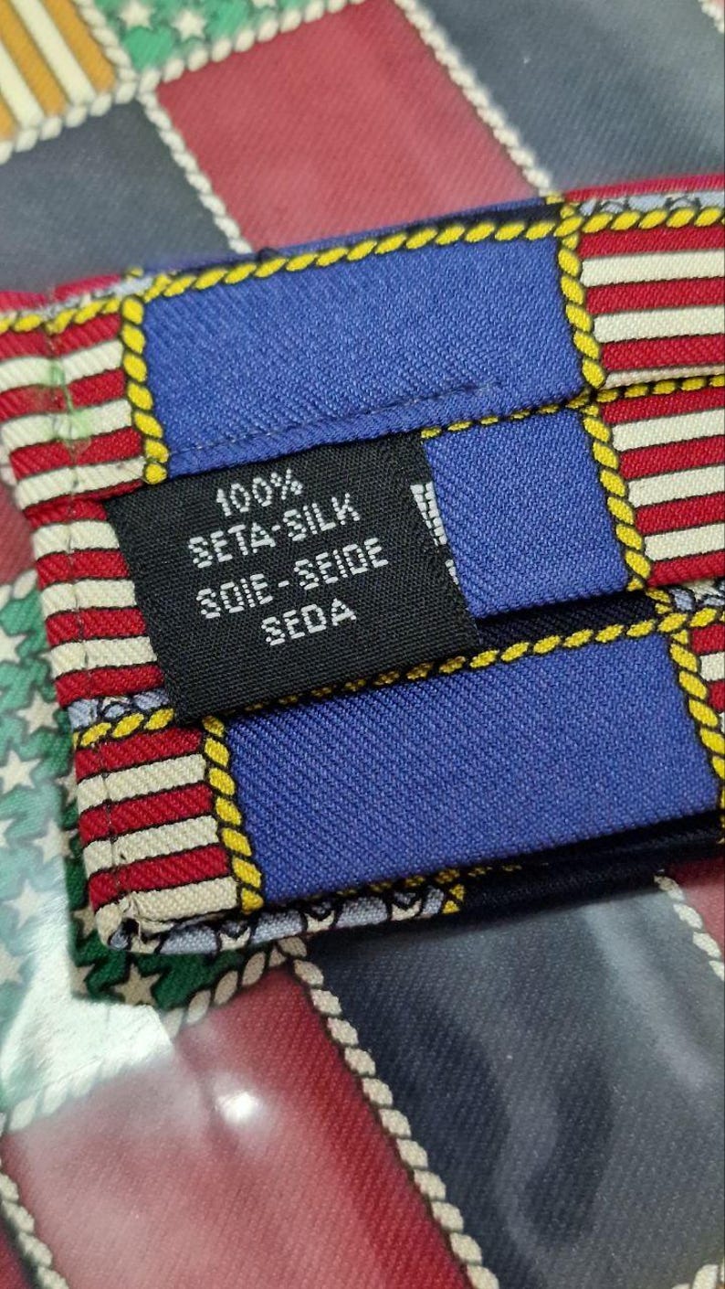 May include: Close-up of a silk tie with a colorful geometric pattern. The tie features blue, red, and white stripes, with yellow rope-like borders. A black label indicates the tie is 100% seta-silk.