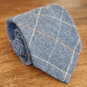 May include: A blue tweed tie with a plaid pattern. The tie features a woven texture with orange, white, and light blue accent lines. The tie is rolled up and resting on a wooden surface.
