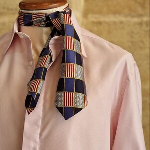 May include: A patterned necktie with a blue, red, and white checkered design is draped over a pink button-down shirt. The tie's pattern includes squares and vertical stripes, creating a classic and stylish look.