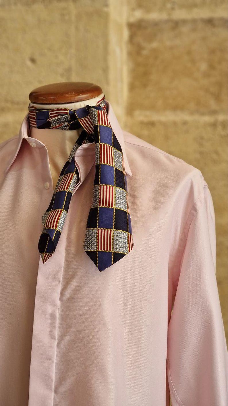 May include: A patterned necktie with a blue, red, and white checkered design is draped over a pink button-down shirt. The tie's pattern includes squares and vertical stripes, creating a classic and stylish look.