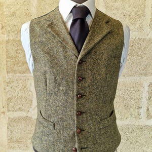 Op de afbeelding: Een tweed vest in olijfgroen en bruin, met een revers, twee voorzakken en een knoopsluiting. Het vest wordt gedragen over een wit overhemd en een donkere stropdas, wat een klassieke, op maat gemaakte look creëert.