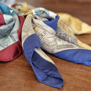 Può includere: Un foulard quadrato in seta con un design multicolore caratterizzato da bordi rosso scuro, blu reale e giallo oro. Il tessuto presenta intricate illustrazioni a linee bianche e nere di architettura urbana e il testo parziale PARIS stampato.