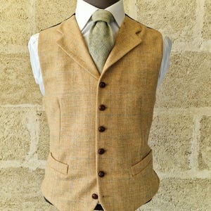 K&ouml;nnte beinhalten: Eine beige Tweedweste mit fallendem Revers, braunen Kn&ouml;pfen und zwei Vordertaschen. Die Weste wird mit einem wei&szlig;en Hemd und einer hellgr&uuml;nen Krawatte kombiniert. Das Outfit wird auf einer Schaufensterpuppe pr&auml;sentiert.