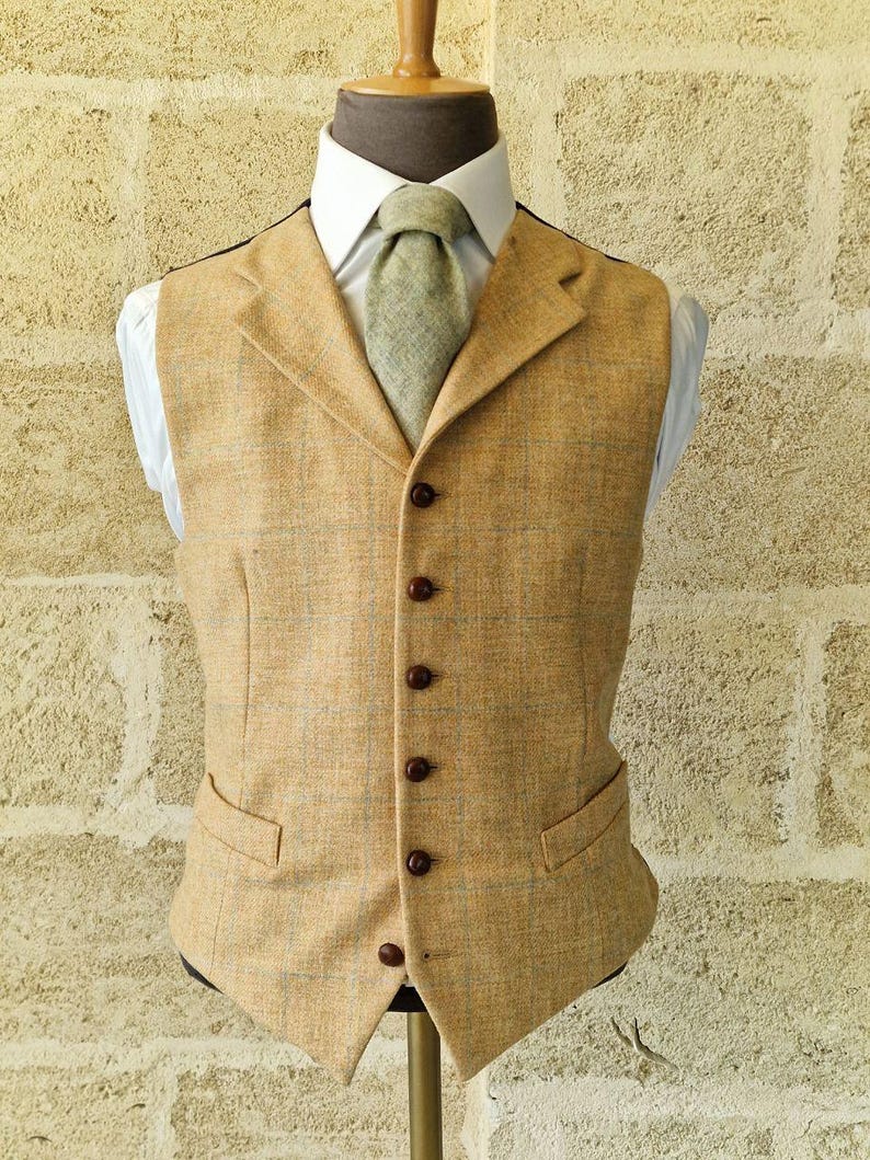 K&ouml;nnte beinhalten: Eine beige Tweedweste mit fallendem Revers, braunen Kn&ouml;pfen und zwei Vordertaschen. Die Weste wird mit einem wei&szlig;en Hemd und einer hellgr&uuml;nen Krawatte kombiniert. Das Outfit wird auf einer Schaufensterpuppe pr&auml;sentiert.