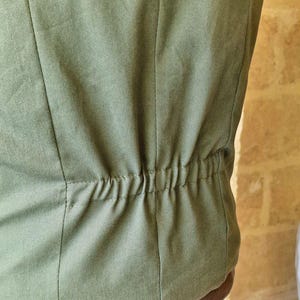 K&ouml;nnte beinhalten: Nahaufnahme eines olivgr&uuml;nen Stoffes, m&ouml;glicherweise ein Kleidungsst&uuml;ck. Vertikale N&auml;hte verlaufen &uuml;ber den Stoff, mit einem gerafften elastischen Detail am unteren Rand. Der Stoff hat ein leicht strukturiertes Aussehen.
