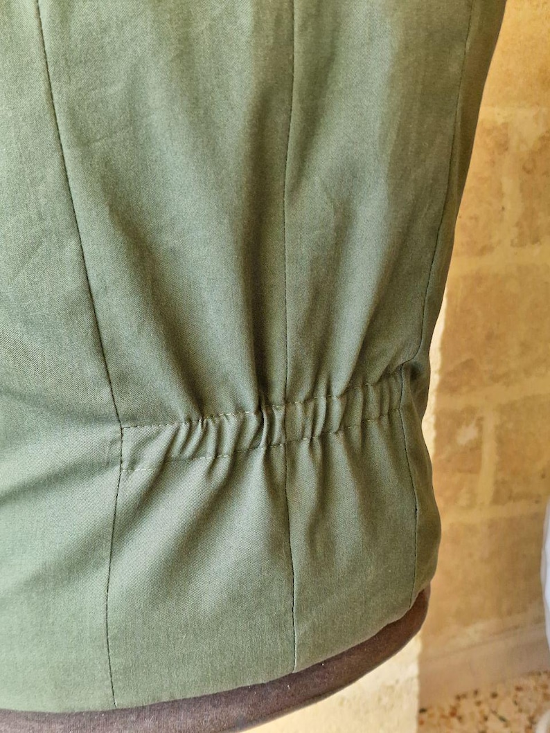 K&ouml;nnte beinhalten: Nahaufnahme eines olivgr&uuml;nen Stoffes, m&ouml;glicherweise ein Kleidungsst&uuml;ck. Vertikale N&auml;hte verlaufen &uuml;ber den Stoff, mit einem gerafften elastischen Detail am unteren Rand. Der Stoff hat ein leicht strukturiertes Aussehen.