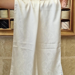 May include: A long, rectangular white scarf featuring a subtle, tonal paisley pattern woven into the fabric. The scarf has a smooth, slightly lustrous finish and a delicate fringe along the bottom edge, with a small white tag attached at the corner.