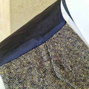 May include: Close-up of a tweed jacket with a navy blue trim and a white collar. The tweed fabric is a mix of beige, navy, and yellow threads. The navy trim borders the tweed and the white collar.