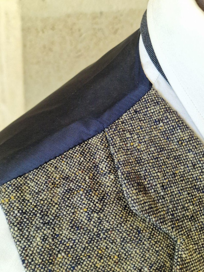 May include: Close-up of a tweed jacket with a navy blue trim and a white collar. The tweed fabric is a mix of beige, navy, and yellow threads. The navy trim borders the tweed and the white collar.