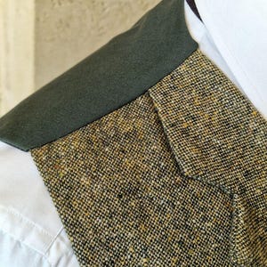 May include: Close-up of a tweed jacket with a dark green collar and a white dress shirt. The tweed fabric is a mix of black, brown, and yellow threads. The collar is a solid dark green color.