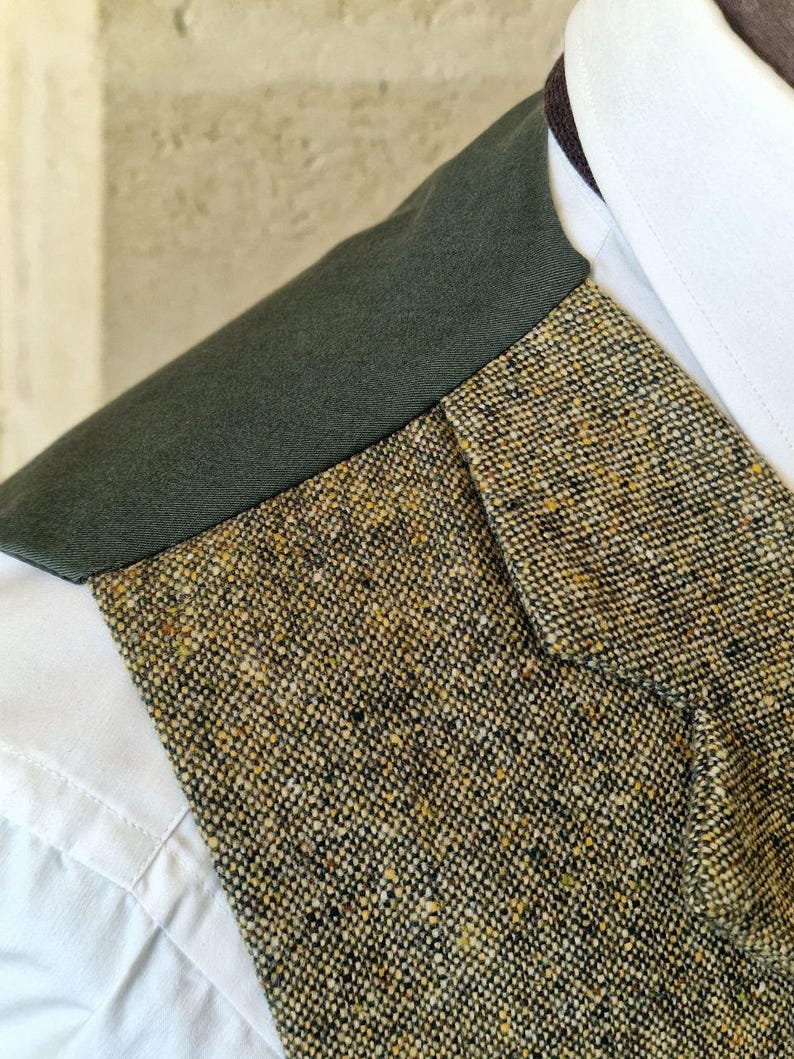 May include: Close-up of a tweed jacket with a dark green collar and a white dress shirt. The tweed fabric is a mix of black, brown, and yellow threads. The collar is a solid dark green color.