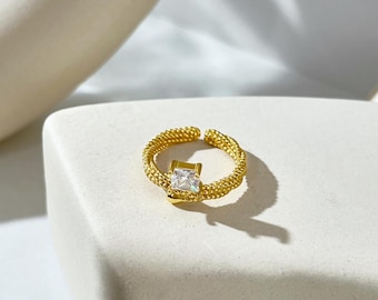 May include: A gold ring with a square-shaped clear stone in the center. The ring is made of a braided metal design.