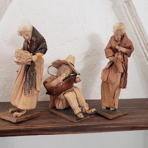 May include: Three handmade corn husk figurines on a wooden shelf. The figures depict people in traditional clothing, including a woman with a basket, a seated musician, and a person holding an object. The figures are on small, dark bases.