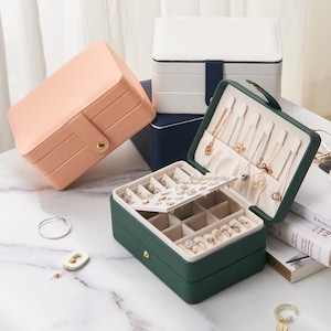 May include: Three jewellery boxes with different coloured lids. The top box is white with a blue trim, the middle box is pink, and the bottom box is green. The green box is open and has compartments for rings, earrings, and necklaces.