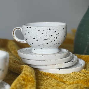Könnte beinhalten: Ein weißes Keramik-Tee-Set mit einem gesprenkelten schwarzen Muster. Die Teetasse hat einen goldfarbenen Henkel und steht auf einem Stapel passender Untertassen. Das Set wird auf einer strukturierten gelben Oberfläche präsentiert.