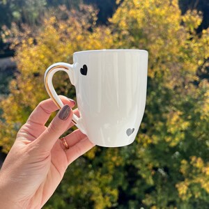 Könnte beinhalten: Weiße Keramiktasse mit goldenem Henkel und Rand, verziert mit drei schwarzen herzförmigen Designs. Die Tasse wird vor einem Hintergrund aus verschwommenem Herbstlaub gehalten.
