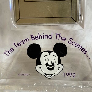 May include: A clear plastic frame with a gold-colored picture frame inside. The frame has a Mickey Mouse image and the text "The Team Behind The Scenes... ©DISNEY 1992".