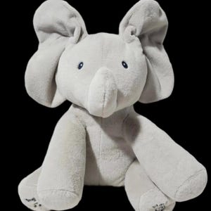 Puede incluir: Un peluche gris con forma de elefante con orejas grandes, una trompa y pequeños ojos azules. El elefante está sentado con los brazos y las piernas extendidos. La palabra "Sing" es visible en un pie.