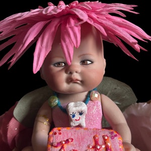 Puede incluir: Una muñeca de porcelana con una corona de flores rosas y un vestido rosa brillante con un dije de diente. La muñeca sostiene un pastel rosa y blanco con la palabra "Alegría" escrita en él.