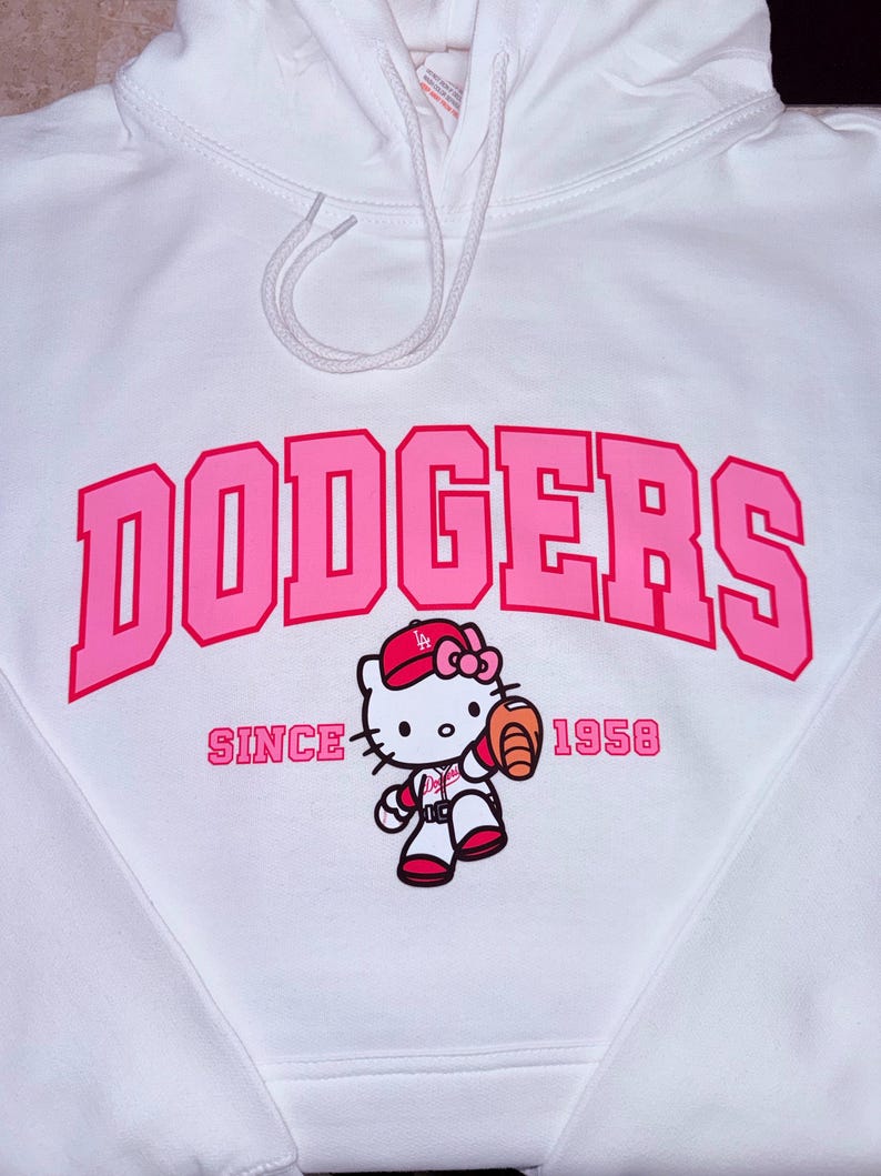May include: White hoodie with a pink "DODGERS" graphic and a baseball-themed Hello Kitty character. The text "SINCE 1958" is also printed on the front. The hoodie has a drawstring hood.