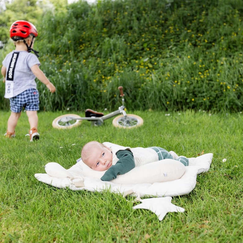 May include: A baby lying on a white star-shaped play mat with a green and white striped blanket. The baby is wearing a green long-sleeved shirt and looking at the camera. A toddler wearing a red helmet and a blue and white plaid shirt is walking away from the camera in the background.