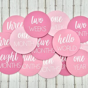 K&ouml;nnte beinhalten: Eine Sammlung runder Baby-Meilensteinkarten in Rosa- und Hellrosa-T&ouml;nen. Jede Karte zeigt wei&szlig;en Text mit Angaben zu Wochen, Monaten und "hello world". Die Karten liegen auf einer hellen Holzmaserung.