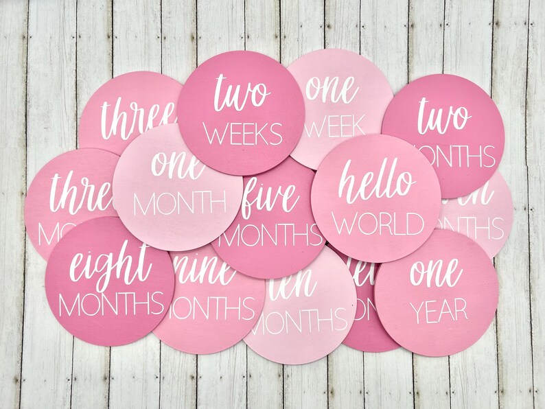 K&ouml;nnte beinhalten: Eine Sammlung runder Baby-Meilensteinkarten in Rosa- und Hellrosa-T&ouml;nen. Jede Karte zeigt wei&szlig;en Text mit Angaben zu Wochen, Monaten und "hello world". Die Karten liegen auf einer hellen Holzmaserung.