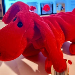 Può includere: Un cane di peluche rosso con occhi neri e naso nero. Il cane è sdraiato sul fianco con la testa girata a sinistra. Il cane ha un'etichetta attaccata alla zampa.