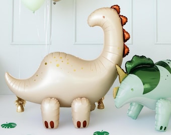 Pode incluir: Um grande balão em forma de dinossauro bege com detalhes dourados e uma crista vermelha, ao lado de um balão triceratops verde e branco. Vários balões flutuam ao fundo, com recortes de folhas verdes espalhados na superfície.