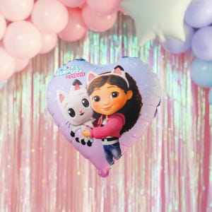 Könnte beinhalten: Herzförmiger Ballon mit Gabby und einem weißen Katzencharakter aus dem Film "Gabby's Dollhouse". Der Ballon hat ein lila-rosa Farbschema mit dem Filmtitel oben. Der Hintergrund umfasst rosa und pastellfarbene Ballons.