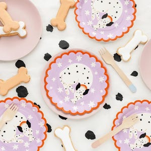 Peut inclure: Une table de fête sur le thème des dalmatiens. Comprend des assiettes lilas avec des bords festonnés orange, des biscuits en forme d'os, des fourchettes en bois et des assiettes roses. Les assiettes présentent un motif de dalmatien endormi avec des étoiles blanches.