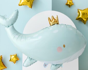 Może przedstawiać: Jasnoniebieski balon w kształcie wieloryba ze złotą koroną, różowymi policzkami i złotymi balonami w kształcie gwiazd. Balon w kształcie wieloryba ma przymocowany sznurek. Tło jest jasnoniebieskie i białe.
