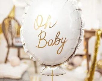 May include: A white, circular foil balloon with the words "Oh Baby" in gold script. The balloon has a frilled edge and is suspended by a gold string. The background is blurred, suggesting a party or celebration.