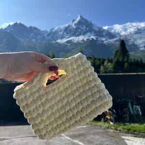 Könnte beinhalten: Eine cremefarbene, handgehäkelte Tragetasche mit rechteckiger Form und Griff. Die Tasche hat ein strukturiertes, gepufftes Stichmuster. Der Hintergrund zeigt eine Bergkette mit schneebedeckten Gipfeln unter einem strahlend blauen Himmel.