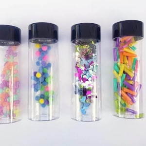 Może przedstawiać: Cztery przezroczyste szklane butelki z czarnymi nakrętkami wypełnione kolorowymi konfetti, brokatem i małymi plastikowymi kształtami. Butelki są ułożone w rzędzie na białym tle.