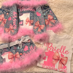 Op de afbeelding: Een set bestaande uit een spijkerjasje, spijkerrokje en wit t-shirt met roze imitatiebont. Het jasje en rokje tonen de naam Brielle, het cijfer 1, roze strikken en het woord Sassy. Het witte t-shirt bevat de tekst Brielle Birthday Girl.