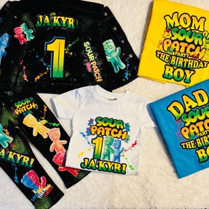 Könnte beinhalten: Ein Sour Patch Kids-thematisches Geburtstags-Outfit-Set für ein einjähriges Kind. Das Set beinhaltet eine schwarze Jeansjacke mit Farbspritzern und Sour Patch Kids-Grafiken, schwarze Jeans mit Sour Patch Kids-Grafiken, ein weißes T-Shirt mit einer Sour Patch Kids-Grafik und der Nummer eins sowie zwei T-Shirts mit Sour Patch Kids-Grafiken und dem Text "Mama von Sour Patch dem Geburtstagskind" und "Papa von Sour Patch dem Geburtstagskind".