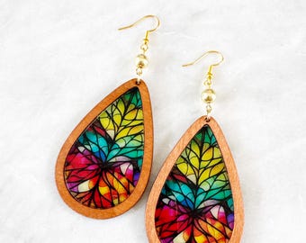 May include: Wooden teardrop earrings with a colorful stained-glass design. The earrings feature a light brown wooden frame with a vibrant, multi-colored floral pattern in the center. Gold-colored ear wires and small beads complete the design.