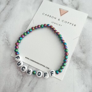 May include: A rainbow iridescent beaded bracelet with white beads spelling out "F U C K O F F". The bracelet is on a white card with the text "CARBON & COPPER JEWELRY" and the website address "www.carbonandcopper.com".