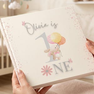 May include: A square hardcover book with a cream-colored cover featuring the text Olivia is 1, a large grey number one, a cartoon bunny holding three pastel balloons, pink butterflies, and a pink flower graphic with speckled pink accents.