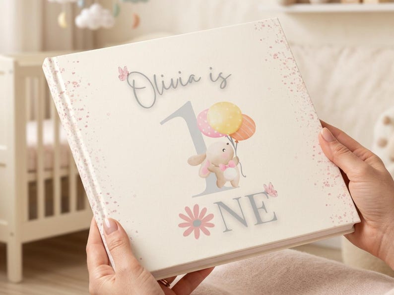 May include: A square hardcover book with a cream-colored cover featuring the text Olivia is 1, a large grey number one, a cartoon bunny holding three pastel balloons, pink butterflies, and a pink flower graphic with speckled pink accents.