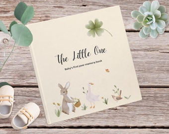 Könnte beinhalten: Ein cremefarbenes Baby-Erinnerungsbuch mit dem Titel "The Little One" und dem Text "Baby's first year memory book". Der Umschlag zeigt eine Aquarell-Illustration eines Kaninchens, einer Gans und eines Vogels.