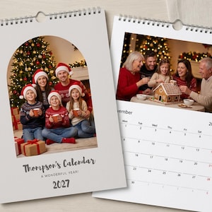 May include: A wall calendar for 2027 with a spiral binding. The cover features an arched photo of a family in holiday sweaters and Santa hats. The interior page shows a November 2027 grid and a photo of a family decorating a gingerbread house.