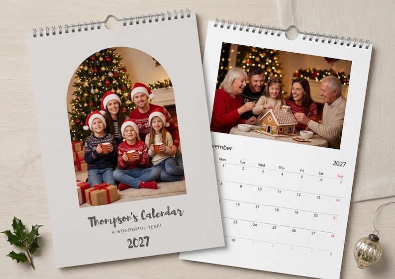 May include: A wall calendar for 2027 with a spiral binding. The cover features an arched photo of a family in holiday sweaters and Santa hats. The interior page shows a November 2027 grid and a photo of a family decorating a gingerbread house.