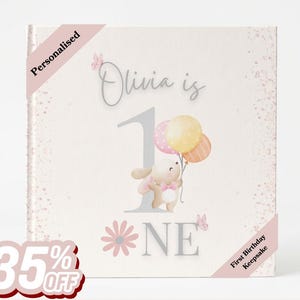 May include: A square book cover with a light cream background featuring a large grey number one, the text Olivia is ONE, a cartoon bunny holding three pastel balloons, pink butterflies, and a pink flower. The cover includes the text Personalised.