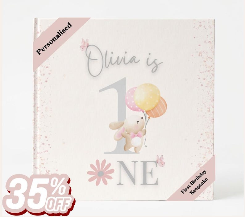 May include: A square book cover with a light cream background featuring a large grey number one, the text Olivia is ONE, a cartoon bunny holding three pastel balloons, pink butterflies, and a pink flower. The cover includes the text Personalised.
