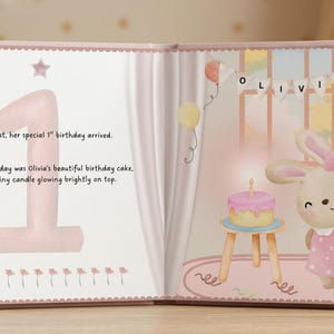 May include: An open children's storybook with pink pages. The left page features a large number one and text reading: And at last, her special 1st birthday arrived. The star of the day was Olivia's beautiful birthday cake, with one tiny candle glowing brightly on top. The right page shows a cartoon rabbit, a cake on a stool, and a banner spelling OLIVIA.
