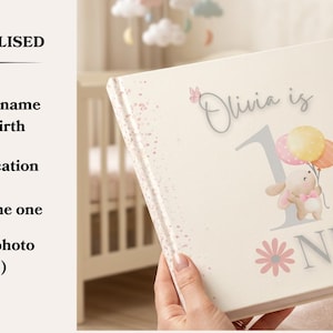 May include: A square photo album with a cream-colored cover featuring the text 'Olivia is 1 NE' in gray and pink. The design includes a watercolor illustration of a bunny holding three pastel balloons, pink butterflies, and a pink flower. The left side lists personalization options including adding a name, date of birth, and dedication message.