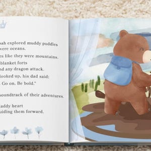 Puede incluir: Un libro infantil abierto con una ilustraci&oacute;n en acuarela de un oso pardo grande y un oso pardo peque&ntilde;o con mochilas azules en un charco de barro. La p&aacute;gina izquierda contiene el texto negro: Daddy Logan and little Noah explored muddy puddles like they were oceans. They climbed park benches like they were mountains. They built blanket forts that could withstand any dragon attack. And every time Noah looked up, his dad said: I'm right here. Go on. Be bold. Little Noah laughter was the soundtrack of their adventures. And his daddy heart was the compass guiding them forward.